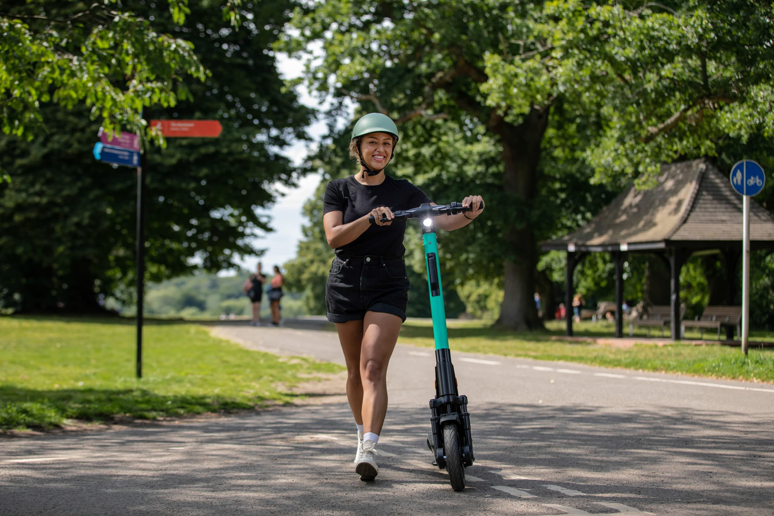 A smiling woman wearing a cycle helmet pushing a Beryl EScooter in a park