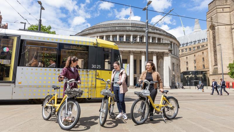 3 young people standing beside yellow Beryl bikes, in front of a tram in Manchester