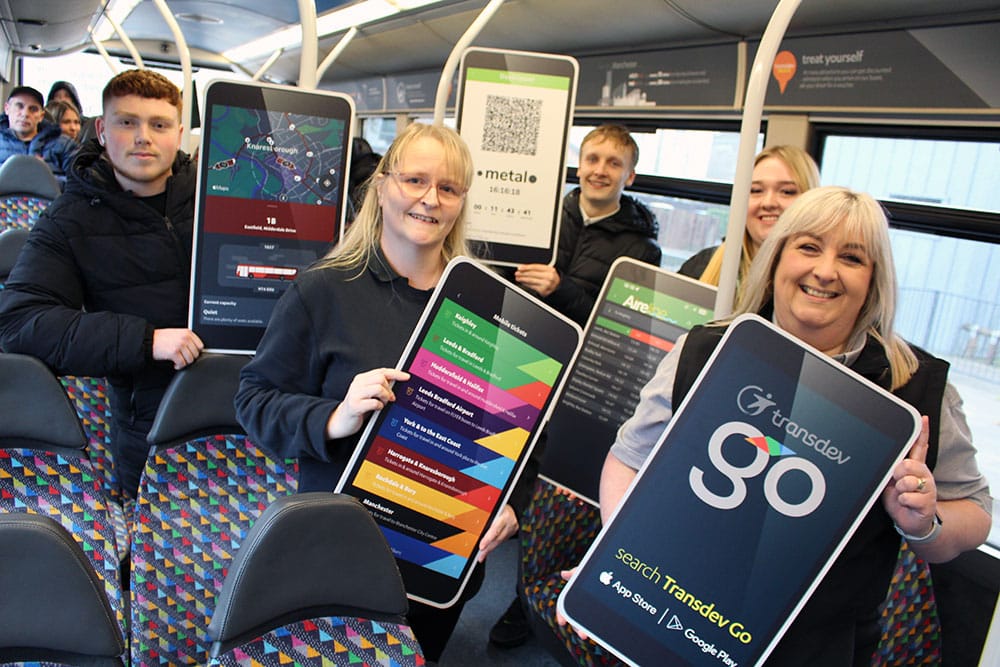 Bus passengers on a Transdev bus holding large images of the Transdev Go app. They are all smiling at the camera.