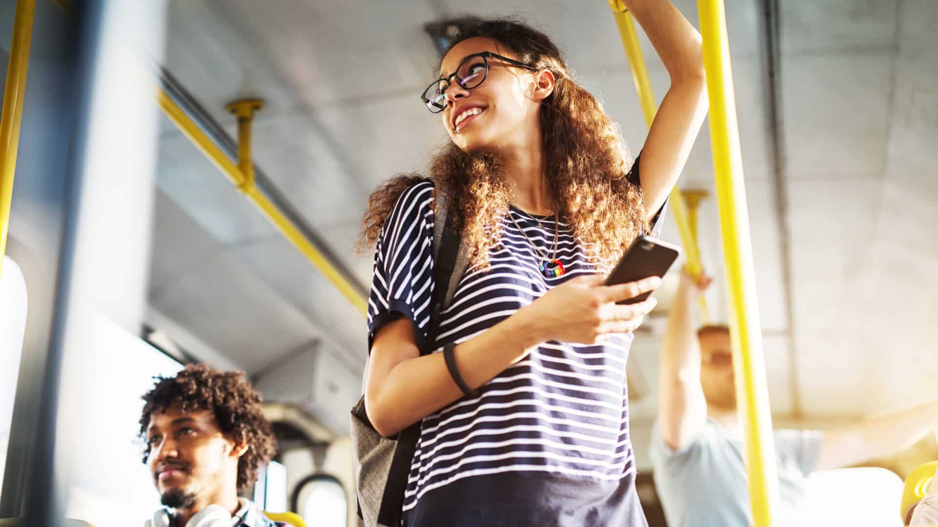 Young woman is standing on the bus using the phone and smiling.