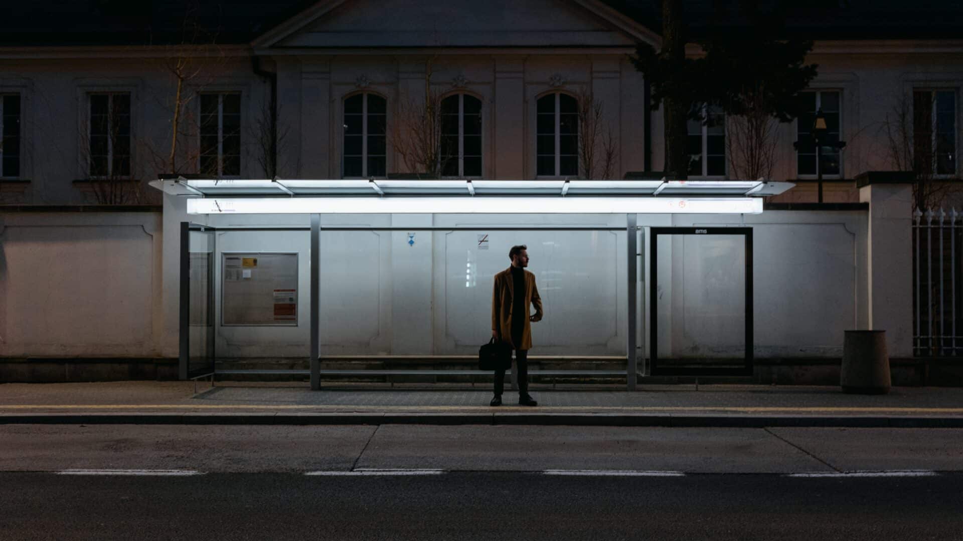 A man standing alone at a bus stop in the dark. The bus stop is some light.