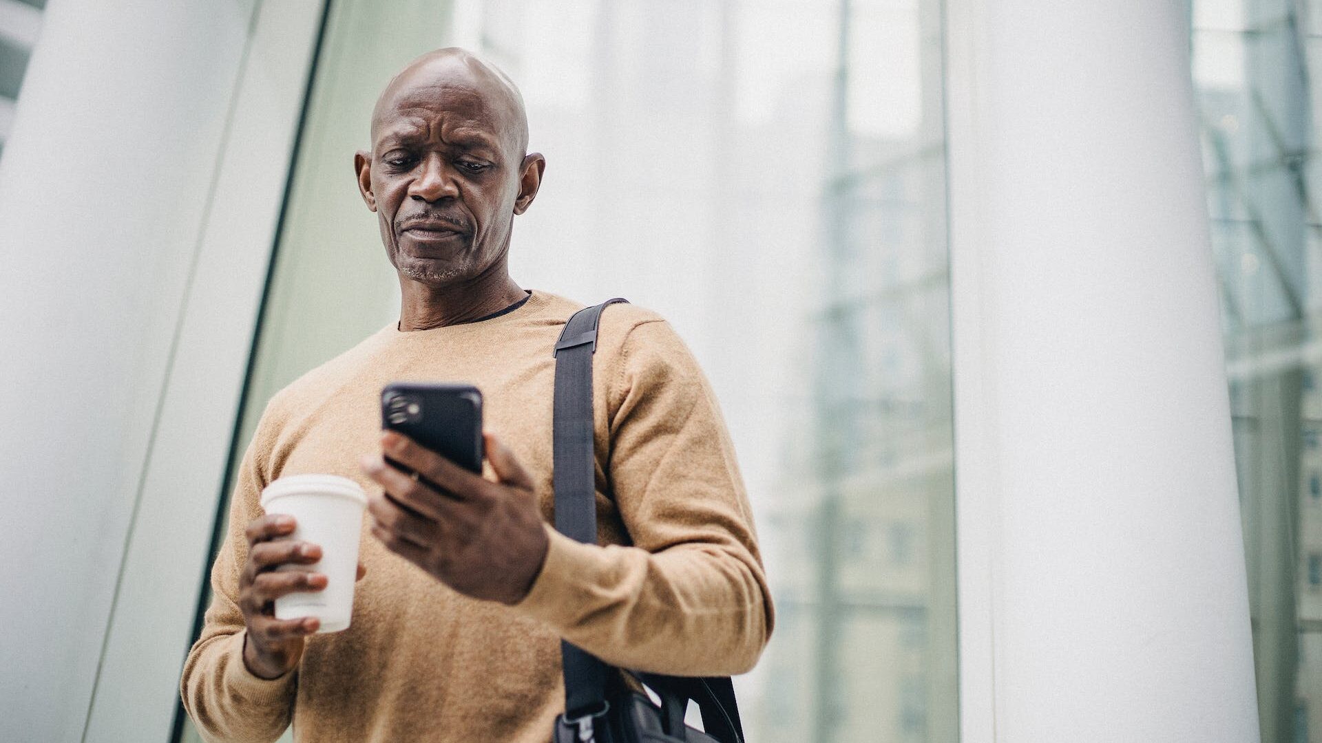 Focused mature black man looking at smartphone holding a coffee on street